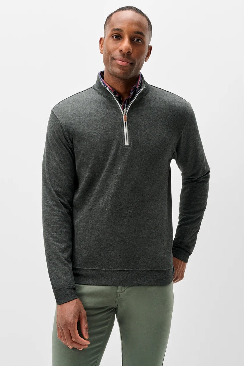 A man wearing a dark gray quarter-zip pullover sweater with a white zipper pull and a patterned shirt collar, paired with olive green pants, against a light gray studio background.