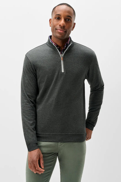 A man wearing a dark gray quarter-zip pullover sweater with a white zipper pull and a patterned shirt collar, paired with olive green pants, against a light gray studio background.