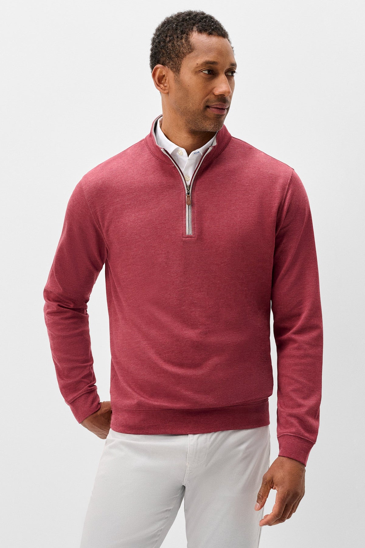 A man wearing a heathered dusty rose quarter-zip pullover sweater over a white collared shirt and white trousers, photographed against a light gray studio background.