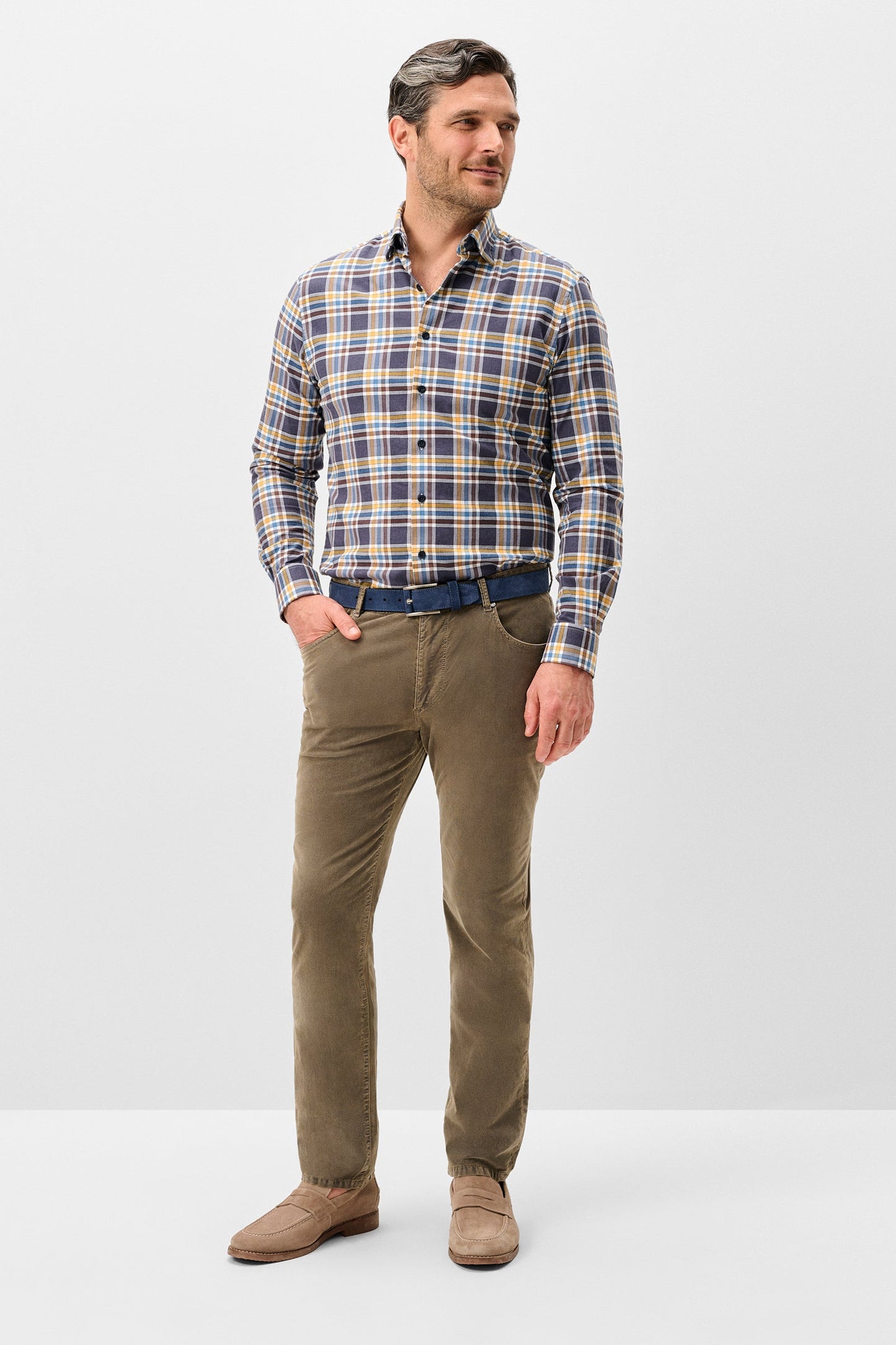 A man modeling a long-sleeve button-up plaid shirt in blue, beige, and white, paired with olive-brown trousers, a dark blue belt, and brown suede shoes, against a light gray studio background.