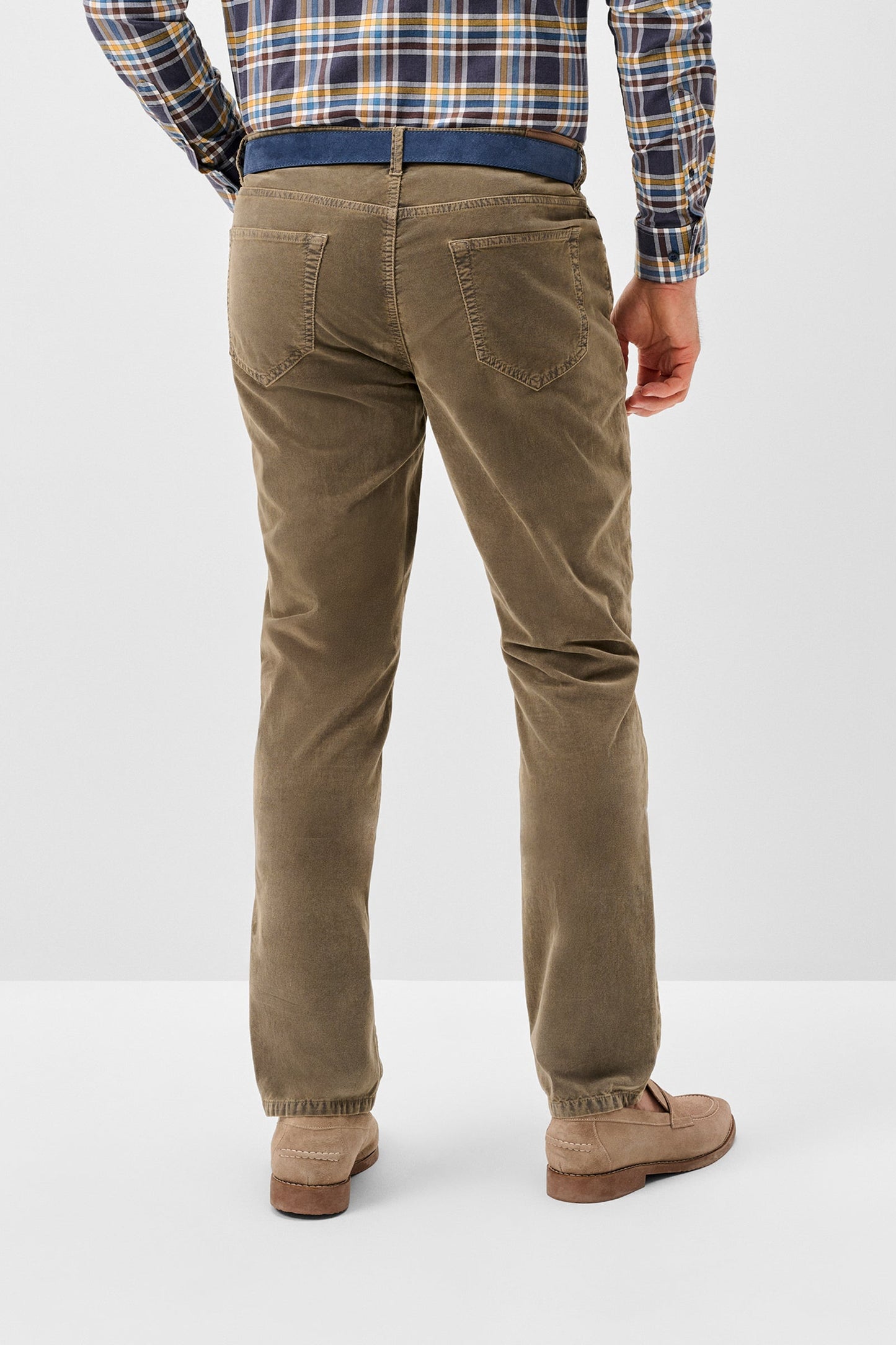 A person shown from the back wearing olive-brown corduroy trousers, a blue belt, a plaid shirt, and tan loafers against a light gray studio background.