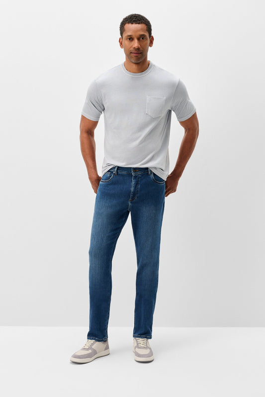 A man modeling a light gray short-sleeve crewneck t-shirt with a chest pocket, paired with blue slim-fit jeans and white sneakers, standing against a plain light gray studio background.