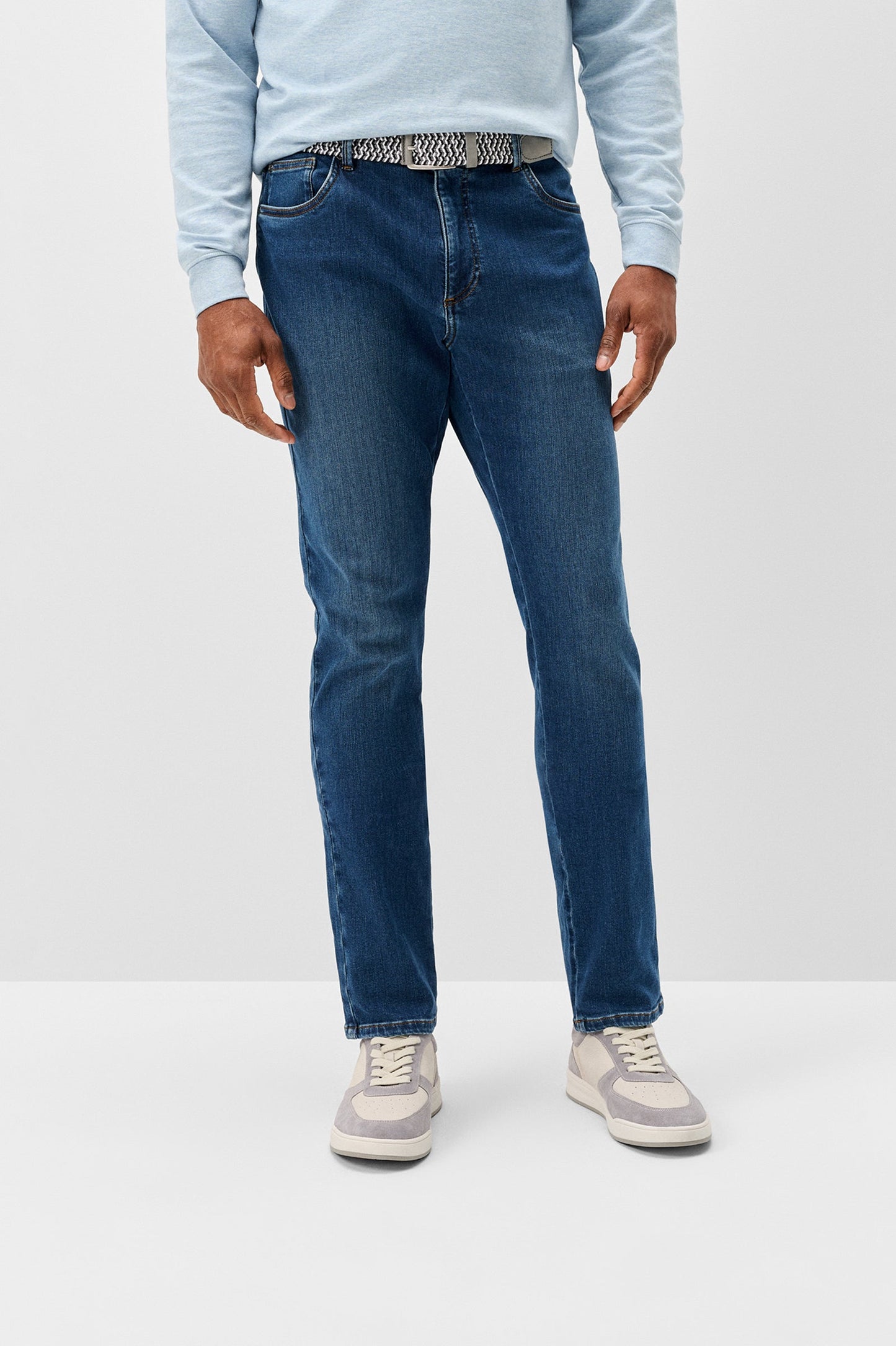 A person wearing medium-wash blue jeans, a light blue long-sleeve top, and gray sneakers, shown from the waist down against a plain light gray studio background.