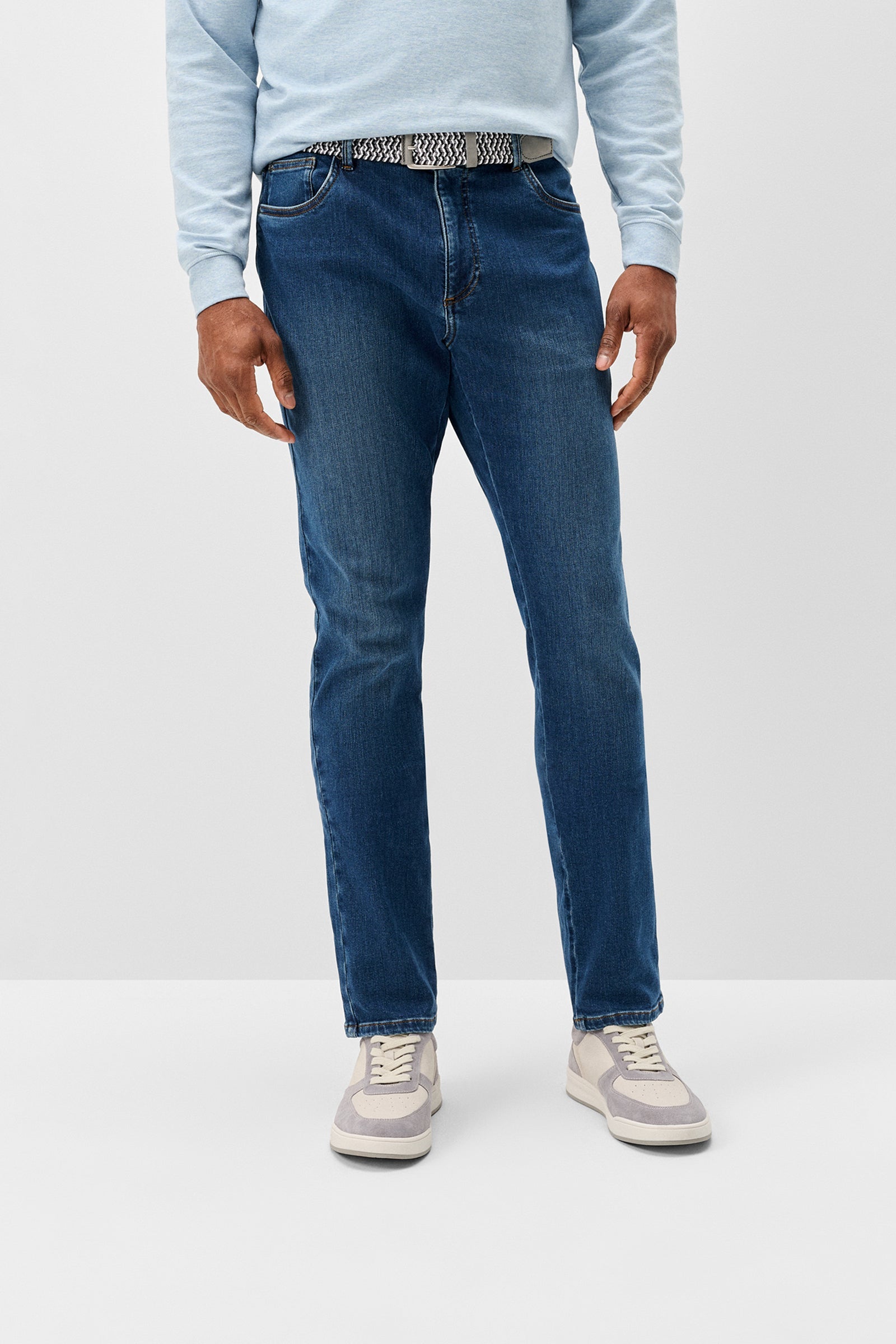 A person wearing medium-wash blue jeans, a light blue long-sleeve top, and gray sneakers, shown from the waist down against a plain light gray studio background.