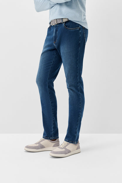 A person wearing dark blue slim-fit jeans with a textured silver belt, a light blue shirt, and beige sneakers, shown from the waist down against a plain white studio background.