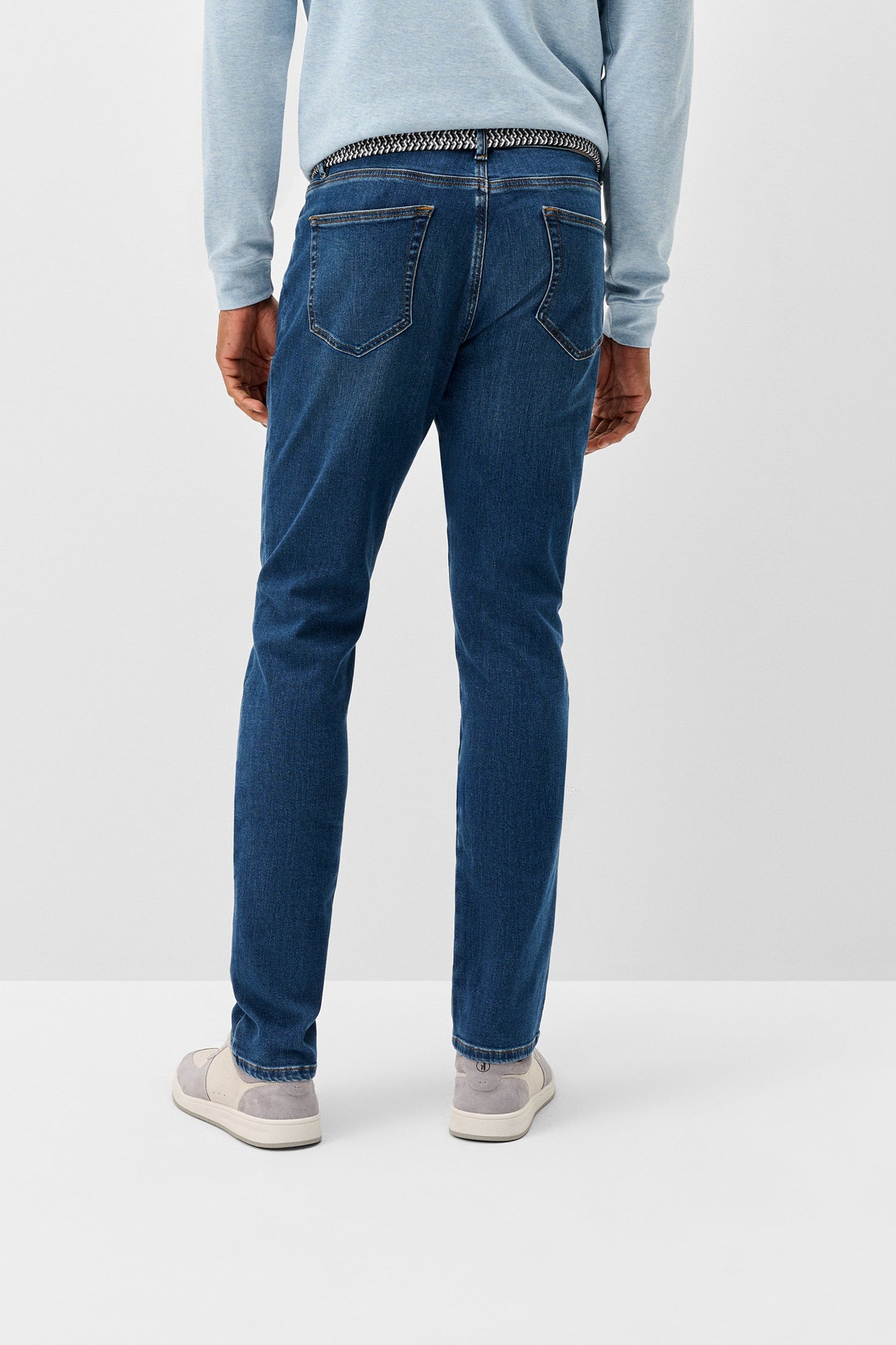 Rear view of a person wearing blue slim-fit jeans, a light blue long-sleeve top, and white sneakers against a plain light gray studio background.