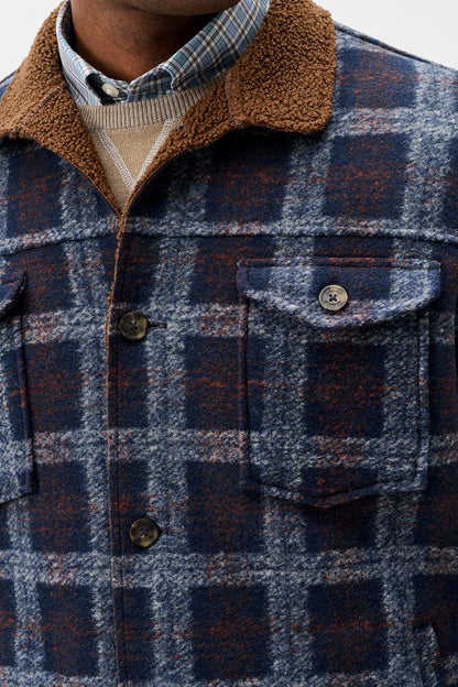 Close-up of a men's plaid overshirt jacket with a brown sherpa fleece collar, worn over a beige knit sweater and blue plaid shirt. The jacket features a blue, gray, and rust-orange check pattern with dark buttons and a chest pocket.