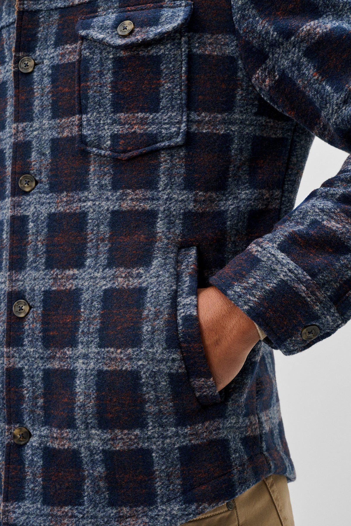 Close-up of a person wearing a navy and blue plaid flannel shirt jacket with brown buttons, showing the chest pocket, front plaid pattern, and hand in pocket.