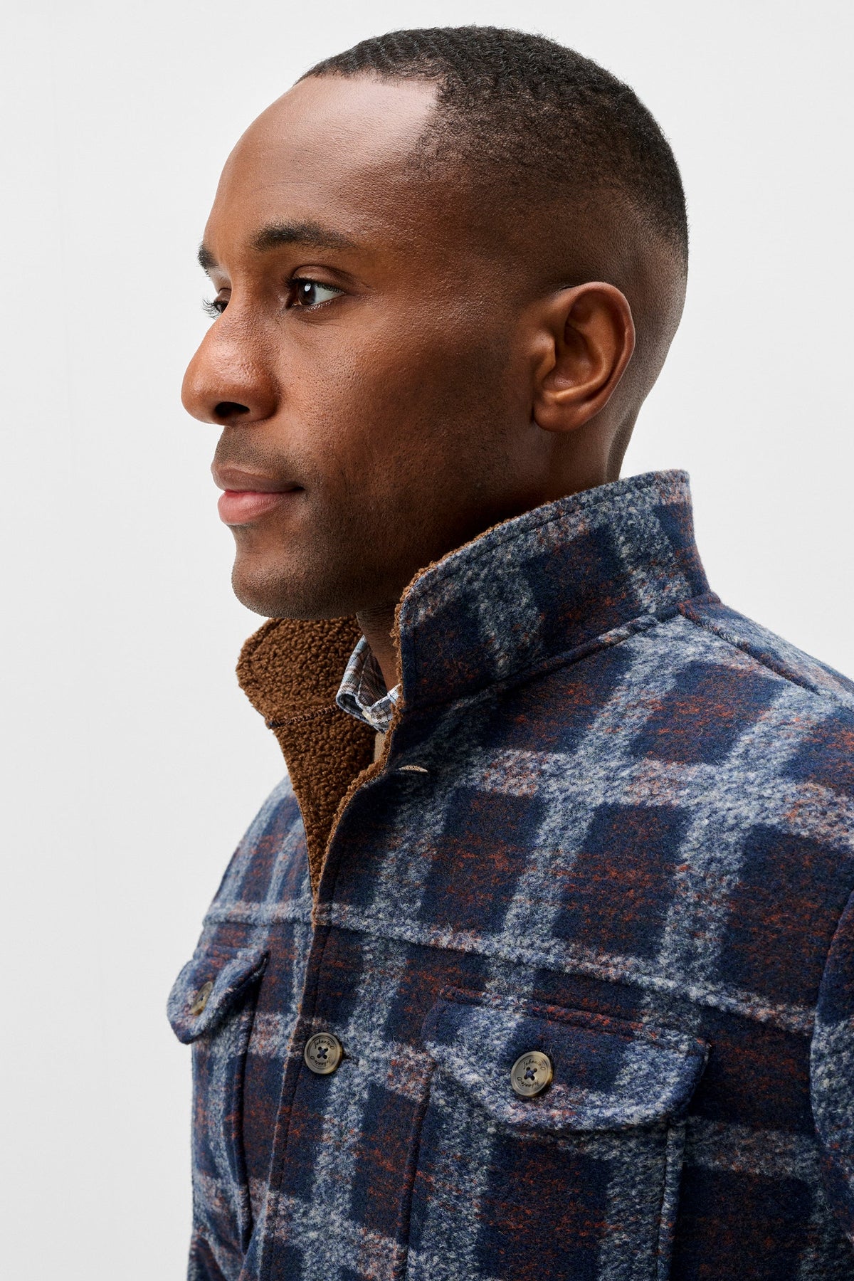 A close-up side profile of a man wearing a blue and brown plaid overshirt with a brown sherpa collar, set against a plain light gray background.