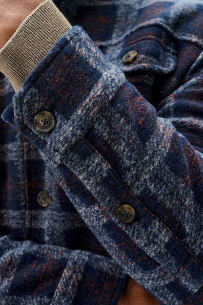 Close-up of a person wearing a textured navy and blue plaid flannel shirt jacket with rust-orange flecks, featuring a ribbed beige cuff and dark buttons.