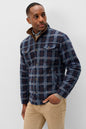A man wearing a navy and blue plaid button-up jacket with a brown collar, paired with beige pants, standing against a light gray studio background.