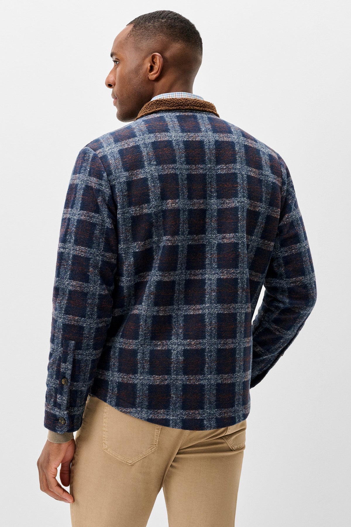 A man shown from the back wearing a navy and blue plaid flannel shirt jacket with a brown corduroy collar, paired with beige trousers, against a light gray studio background.