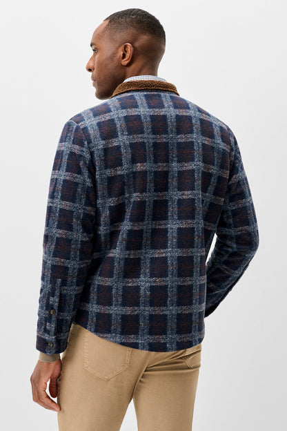 A man shown from the back wearing a navy and blue plaid flannel shirt jacket with a brown corduroy collar, paired with beige trousers, against a light gray studio background.