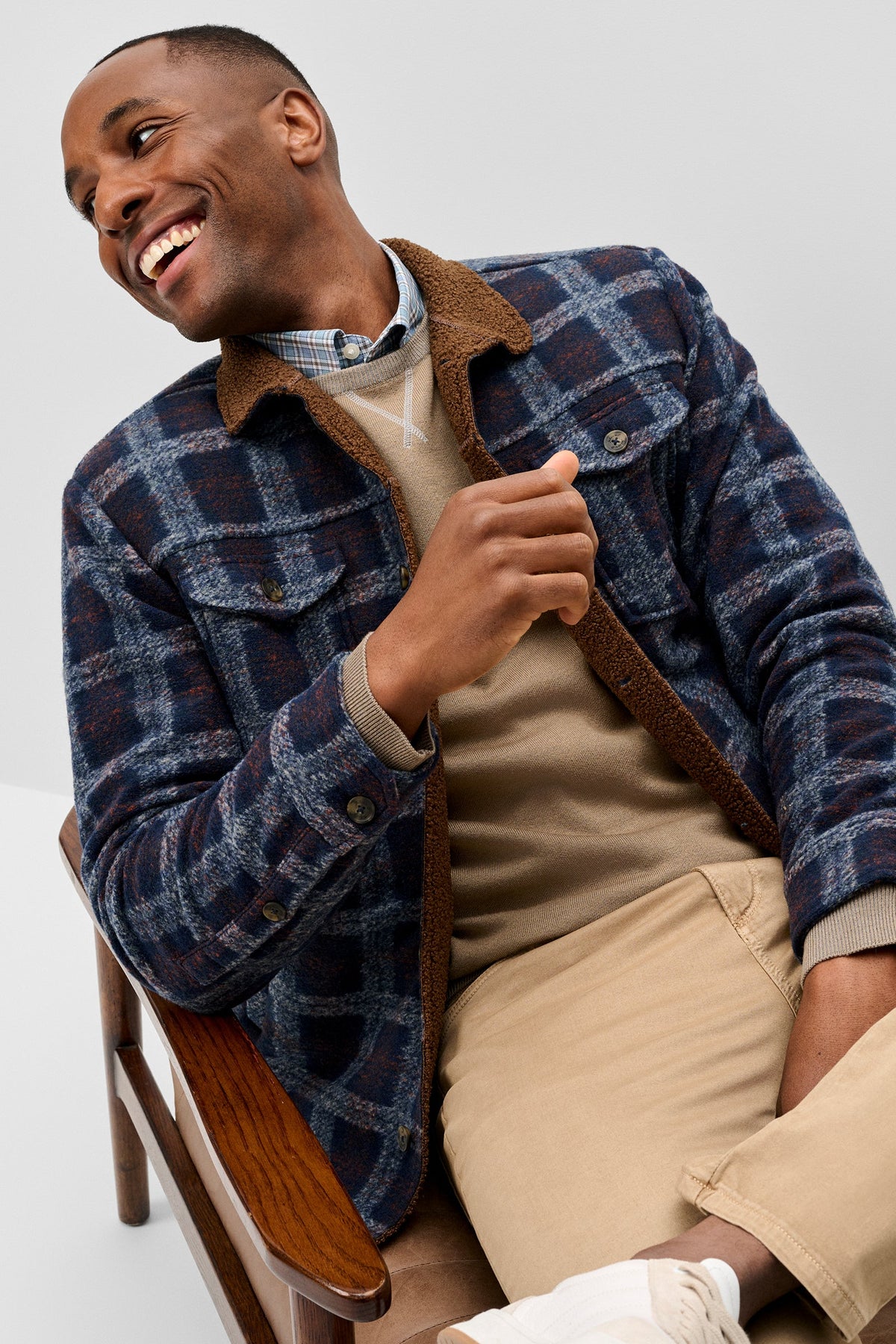 A man smiling while wearing a navy and blue plaid overshirt with brown corduroy collar, layered over a beige sweater and khaki pants, seated on a wooden chair against a light gray studio background.