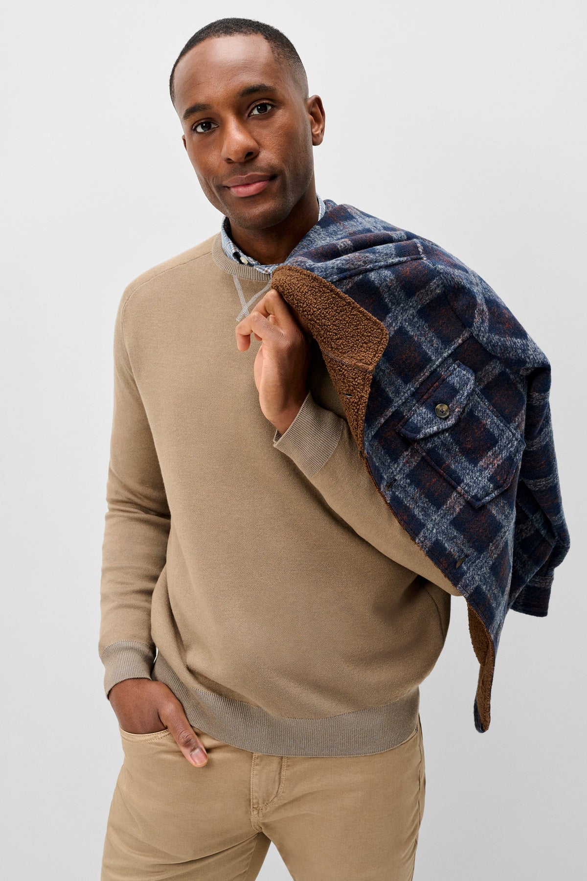 A man wearing a beige crewneck sweater and khaki pants, holding a navy blue plaid flannel shirt with a brown sherpa lining over his shoulder, against a light gray studio background.