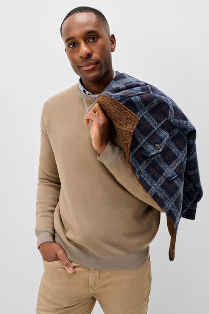 A man wearing a beige crewneck sweater and khaki pants, holding a navy blue plaid flannel shirt with a brown sherpa lining over his shoulder, against a light gray studio background.