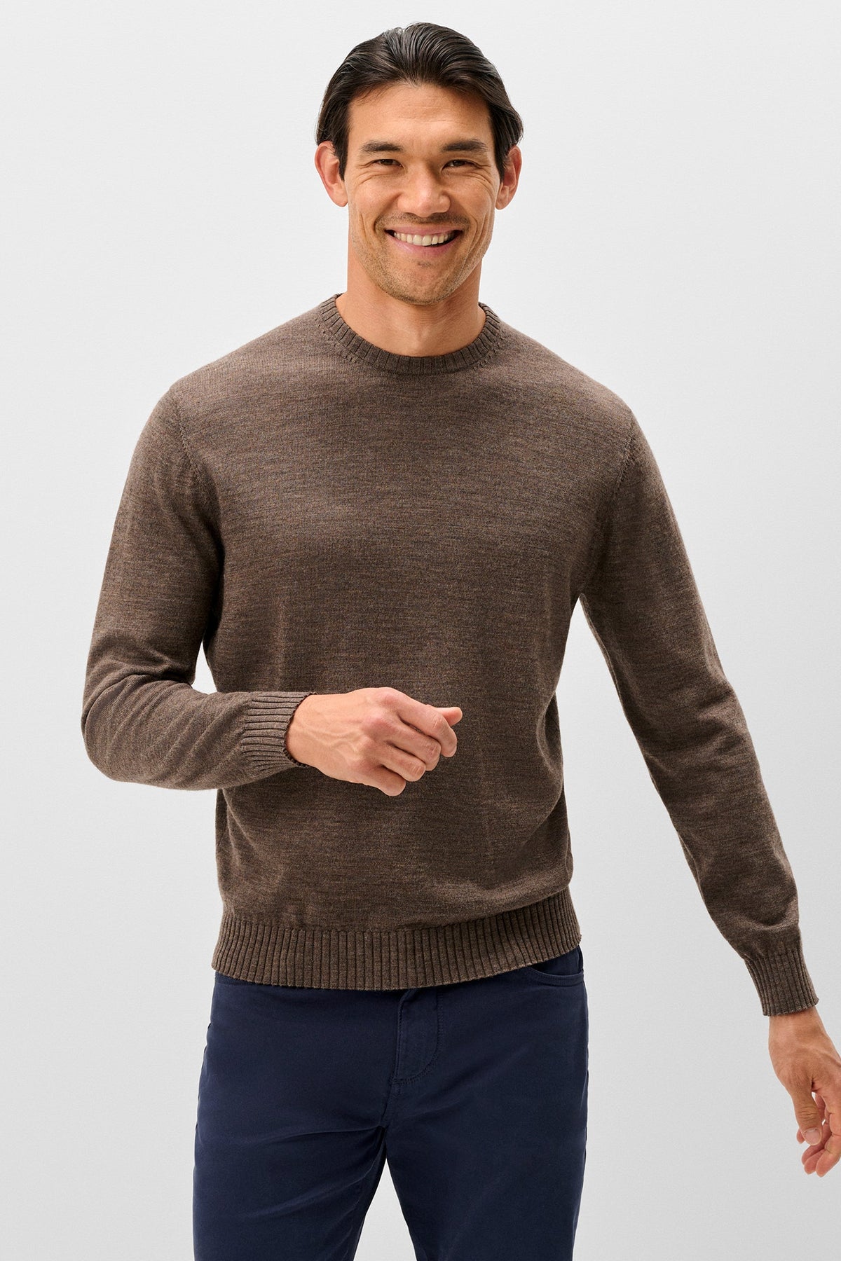 A smiling man wearing a brown long-sleeve knit sweater and navy pants, standing against a light gray studio background.