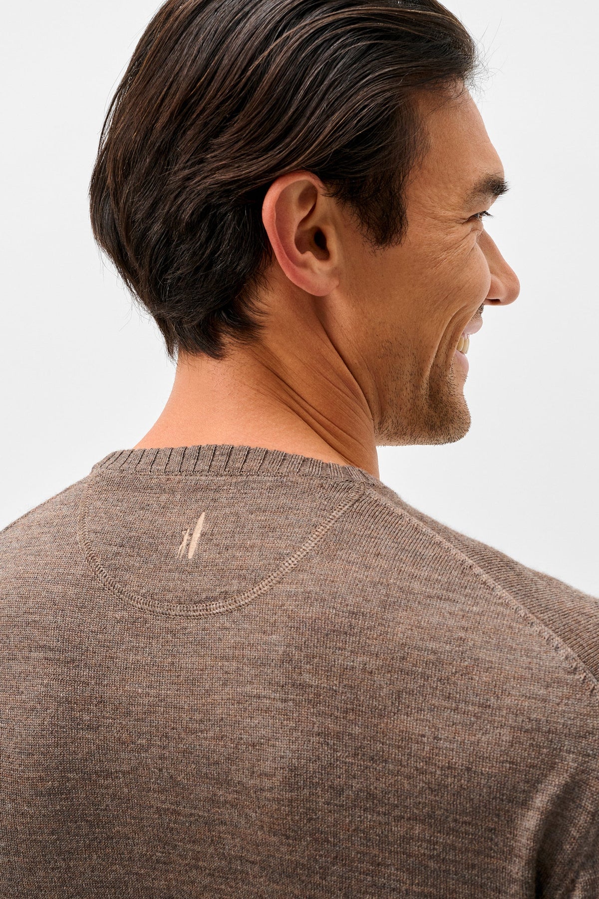 A close-up side profile of a man wearing a brown knit crewneck sweater against a light gray background.