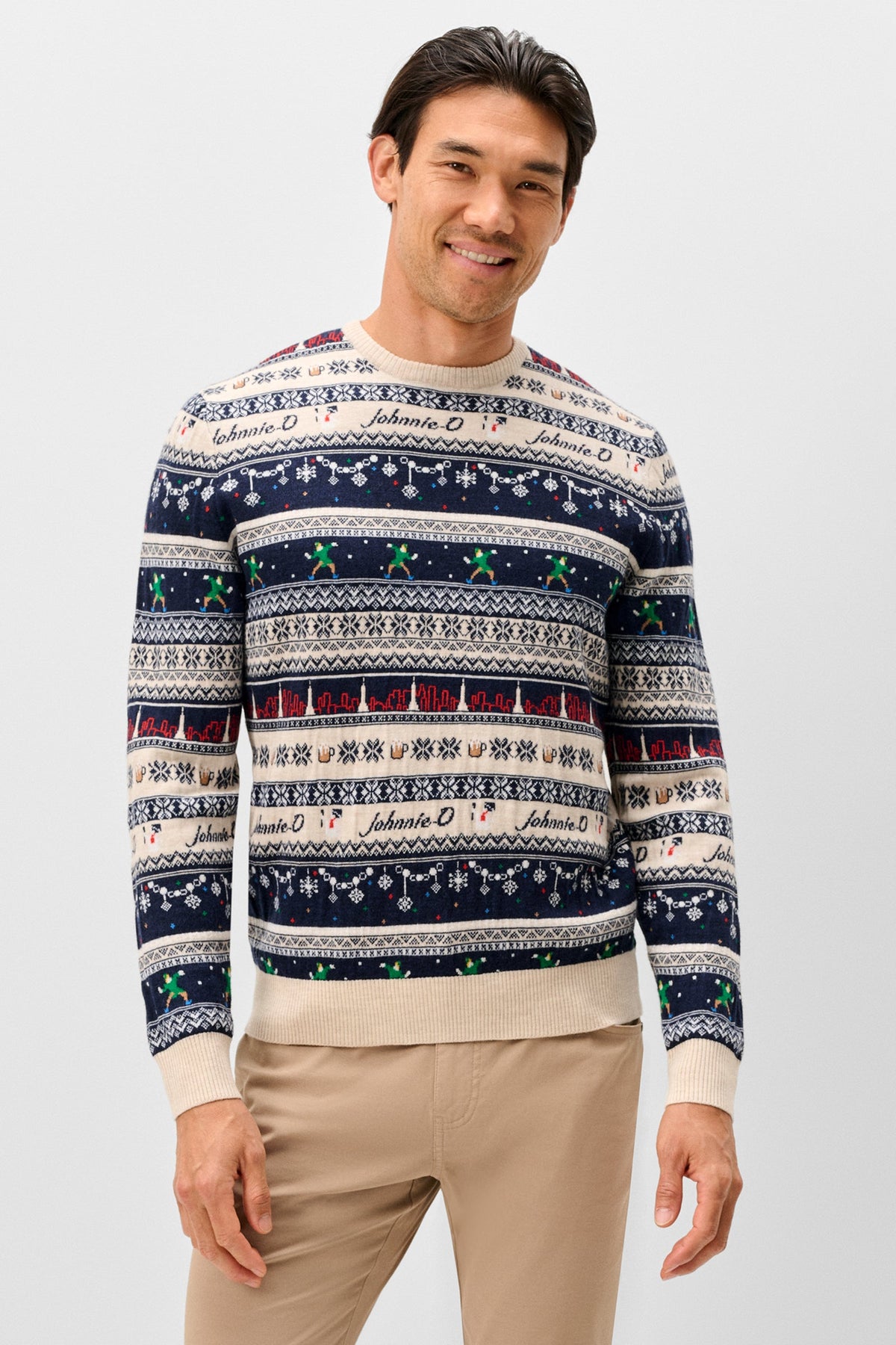 A man wearing a long-sleeve crewneck sweater with a festive Nordic pattern in navy blue, cream, and red, featuring the name 'Johnnie O' repeated across the design. He is also wearing beige trousers, standing against a solid light gray background.