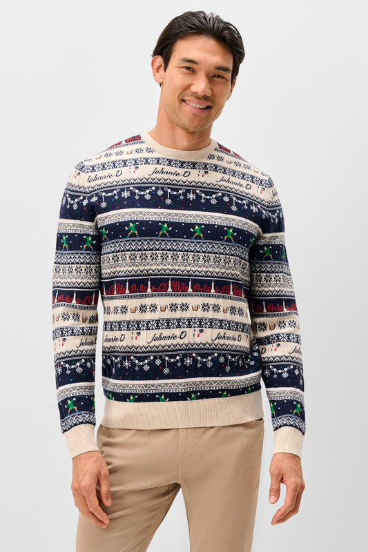 A man wearing a long-sleeve crewneck sweater with a festive Nordic pattern in navy blue, cream, and red, featuring the name 'Johnnie O' repeated across the design. He is also wearing beige trousers, standing against a solid light gray background.