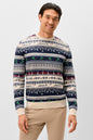 A man wearing a long-sleeve crewneck sweater with a festive Nordic pattern in navy blue, cream, and red, featuring the name 'Johnnie O' repeated across the design. He is also wearing beige trousers, standing against a solid light gray background.