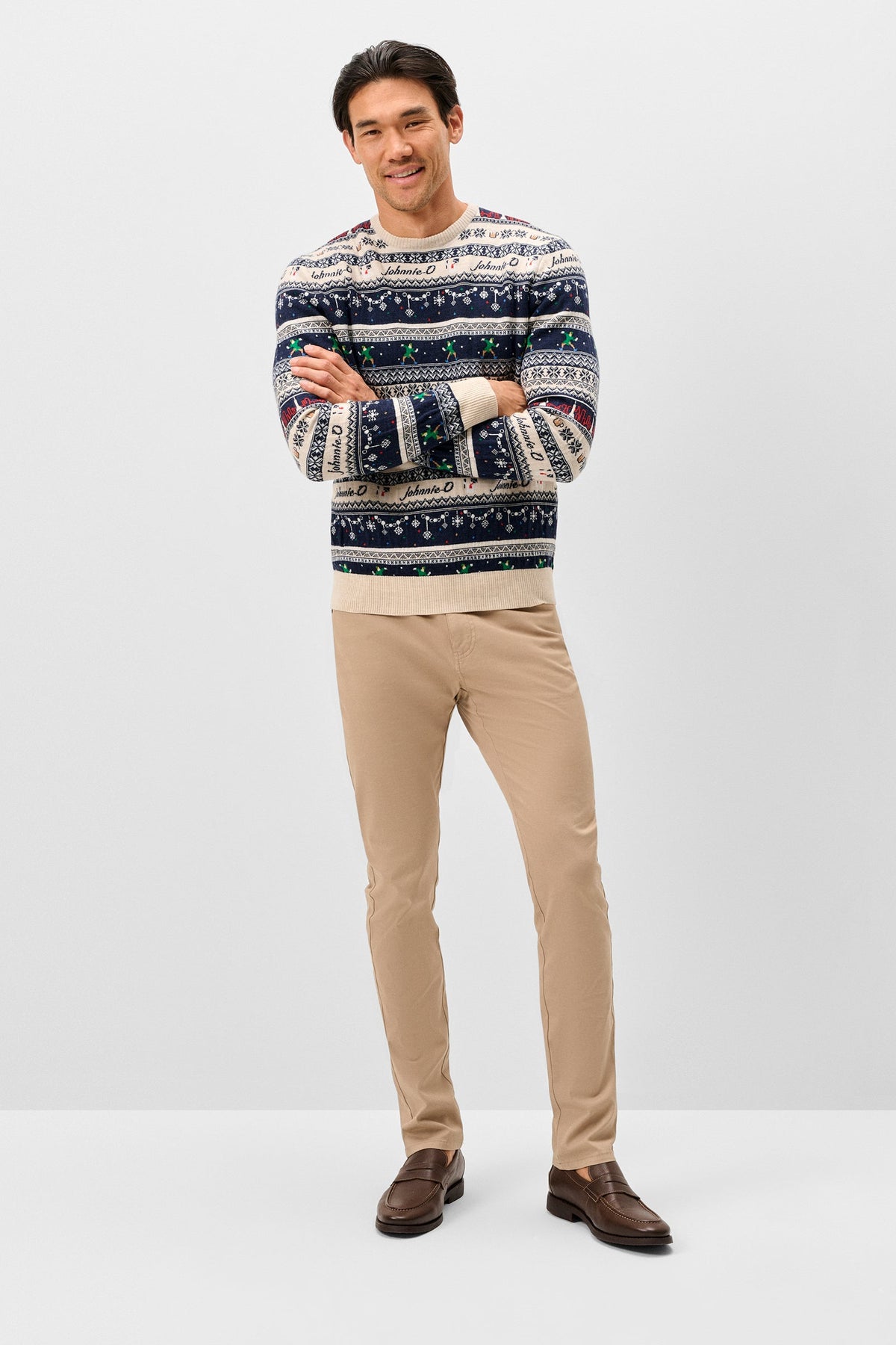 A man modeling a long-sleeve crewneck sweater with a multicolor Nordic/Fair Isle pattern, paired with beige trousers and brown leather shoes, against a light gray studio background.