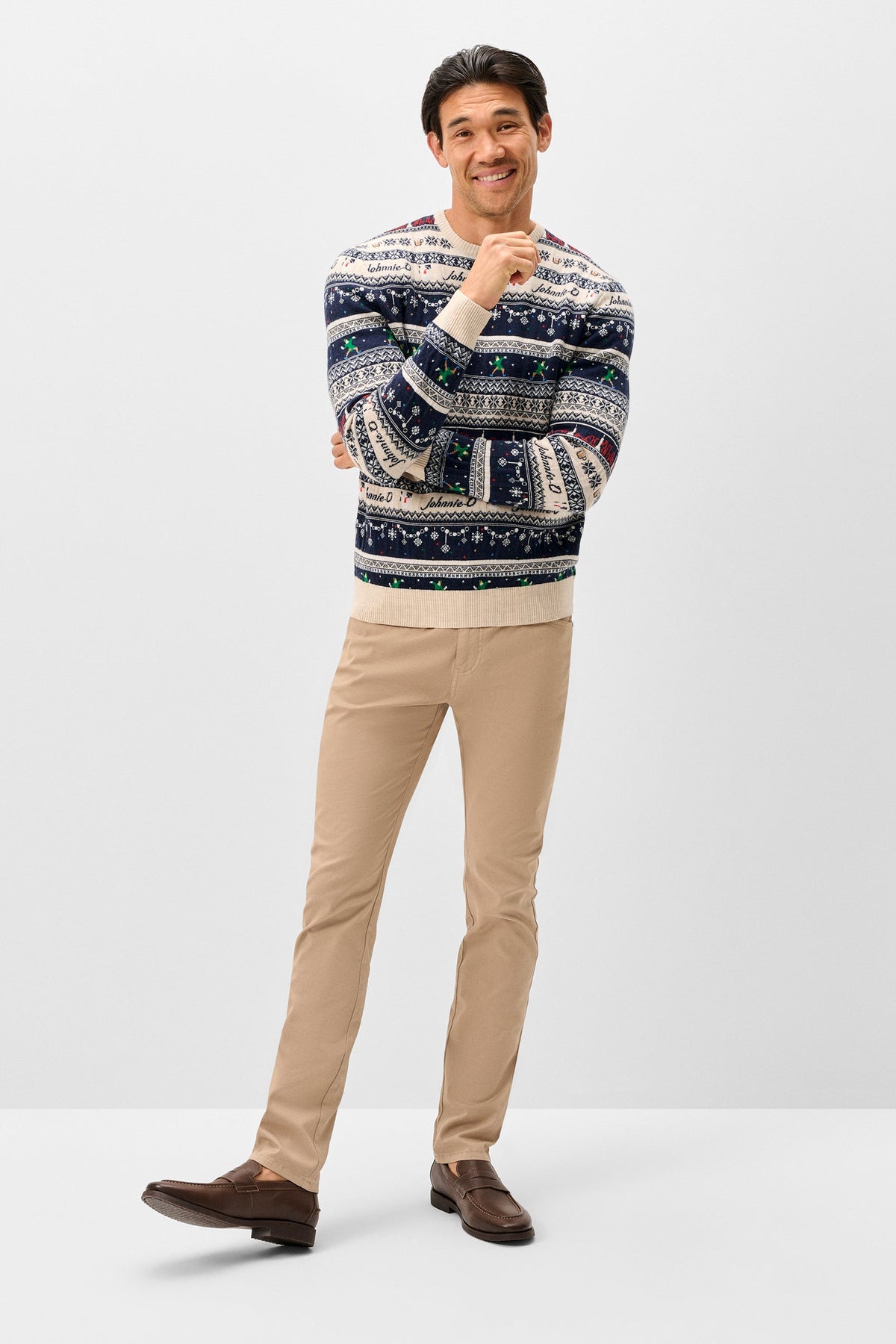 A man modeling a patterned knit sweater with beige trousers and brown loafers, standing against a light gray studio background.