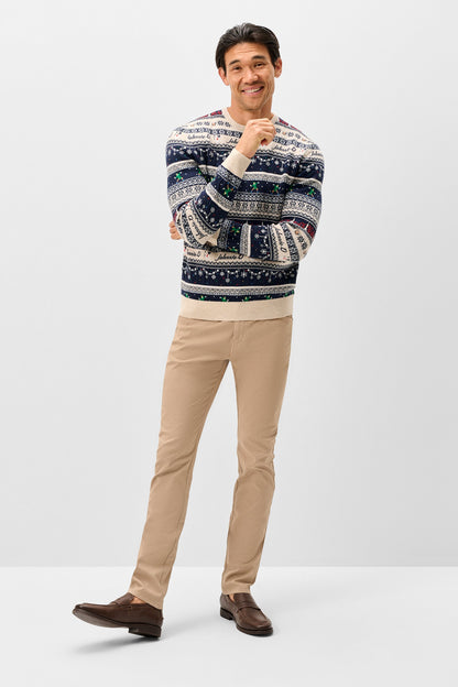 A man modeling a patterned knit sweater with beige trousers and brown loafers, standing against a light gray studio background.