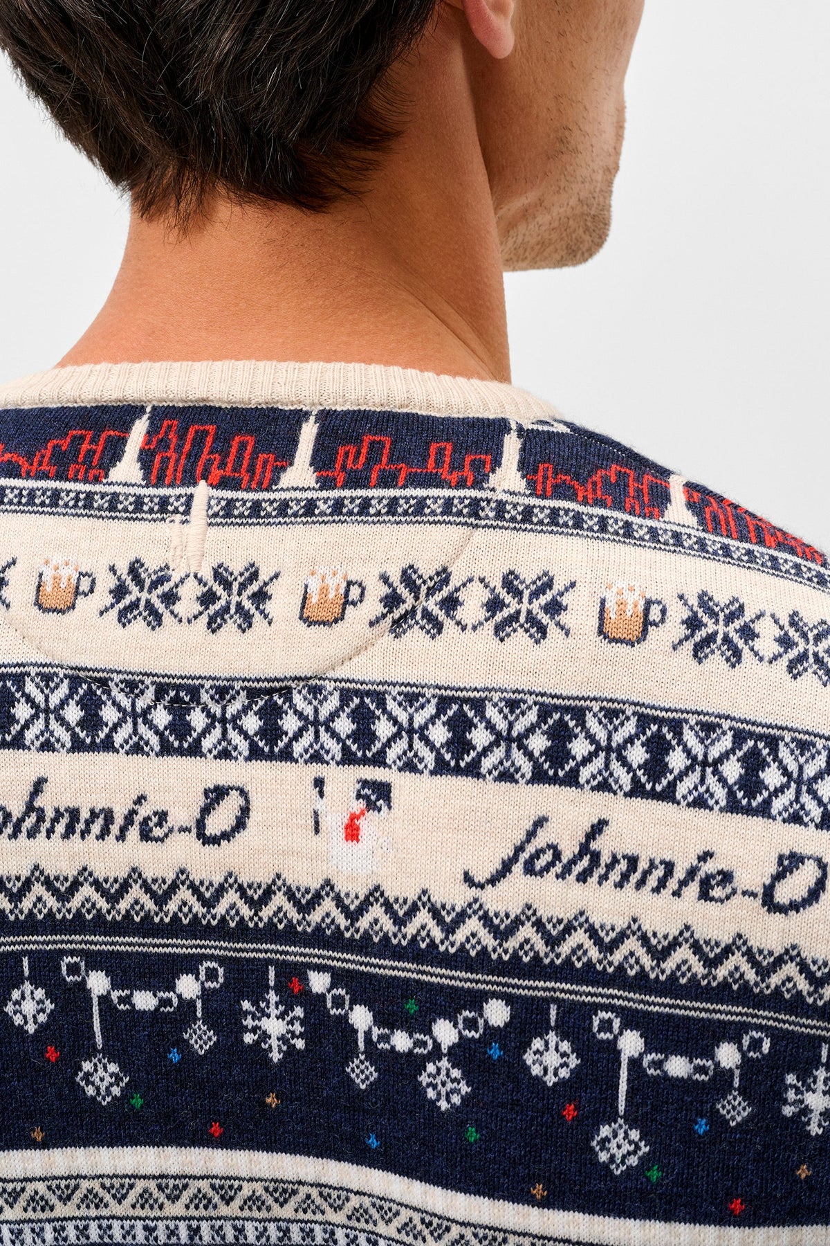 Close-up back view of a man wearing a knitted Christmas sweater with navy, cream, and red patterns, featuring the text 'Johnnie-O' across the back.