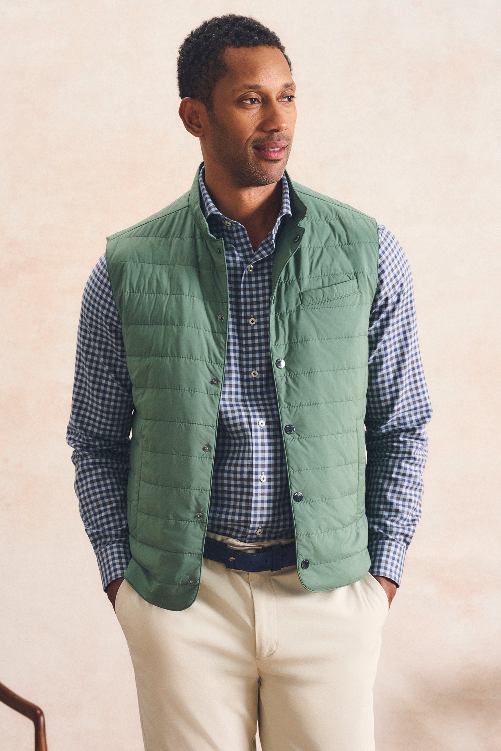 A man wearing a sage green quilted puffer vest over a blue and white checkered shirt, paired with beige trousers, standing against a textured beige wall.