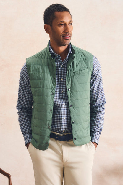 A man wearing a sage green quilted puffer vest over a blue and white checkered shirt, paired with beige trousers, standing against a textured beige wall.