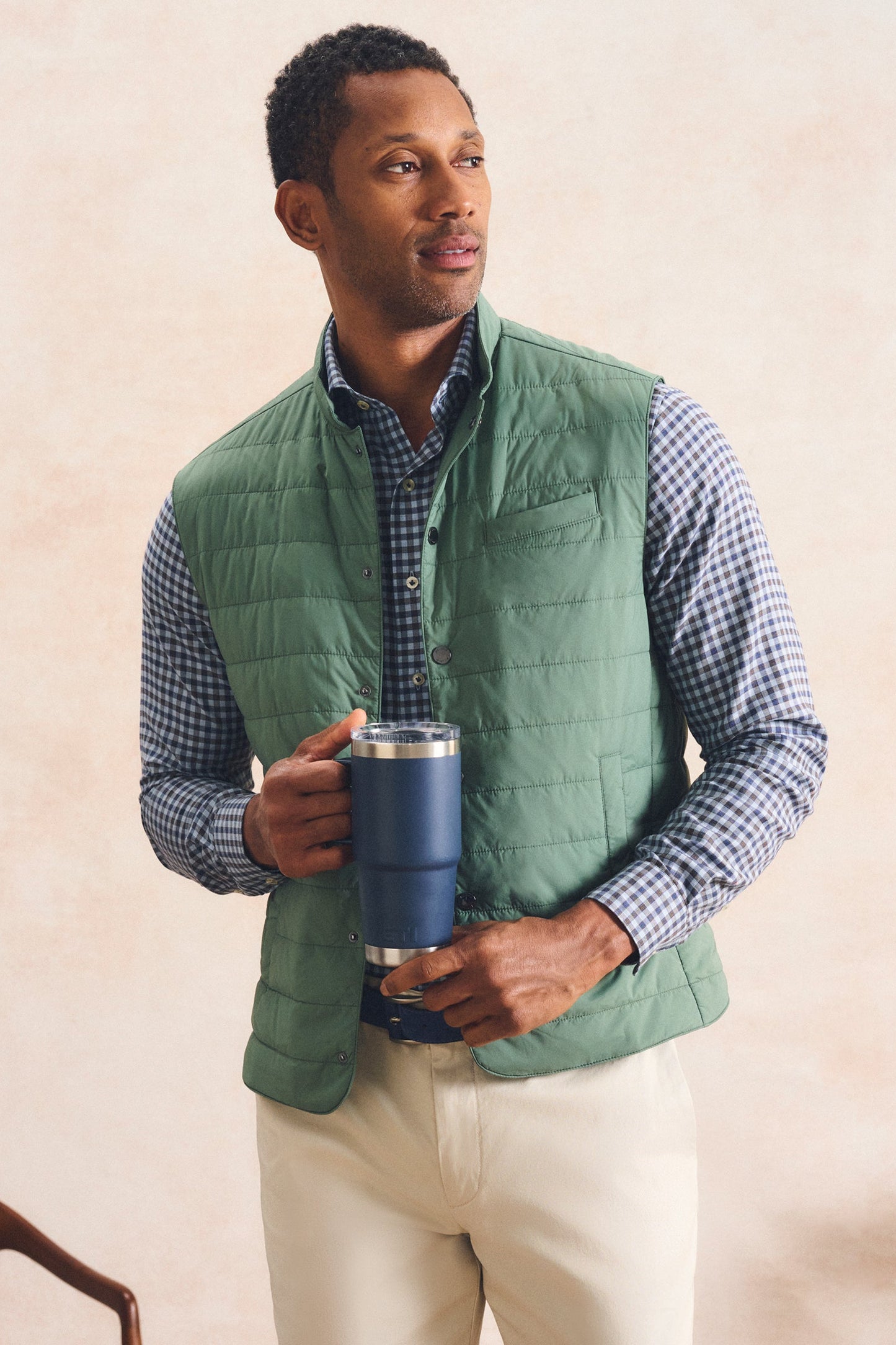 A man wearing a sage green quilted puffer vest over a blue and white checkered shirt, holding a navy blue insulated travel tumbler against a warm beige textured background.