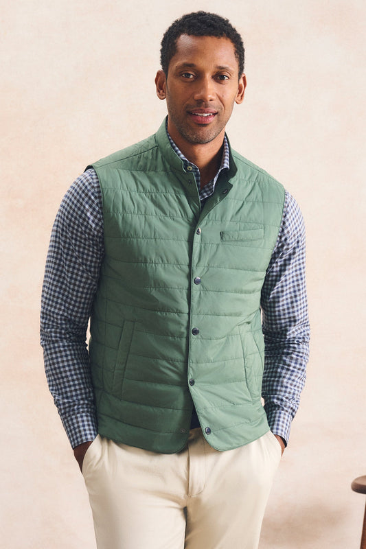 A man wearing a sage green quilted puffer vest over a blue and white checkered shirt, paired with cream-colored pants, standing against a textured beige background.