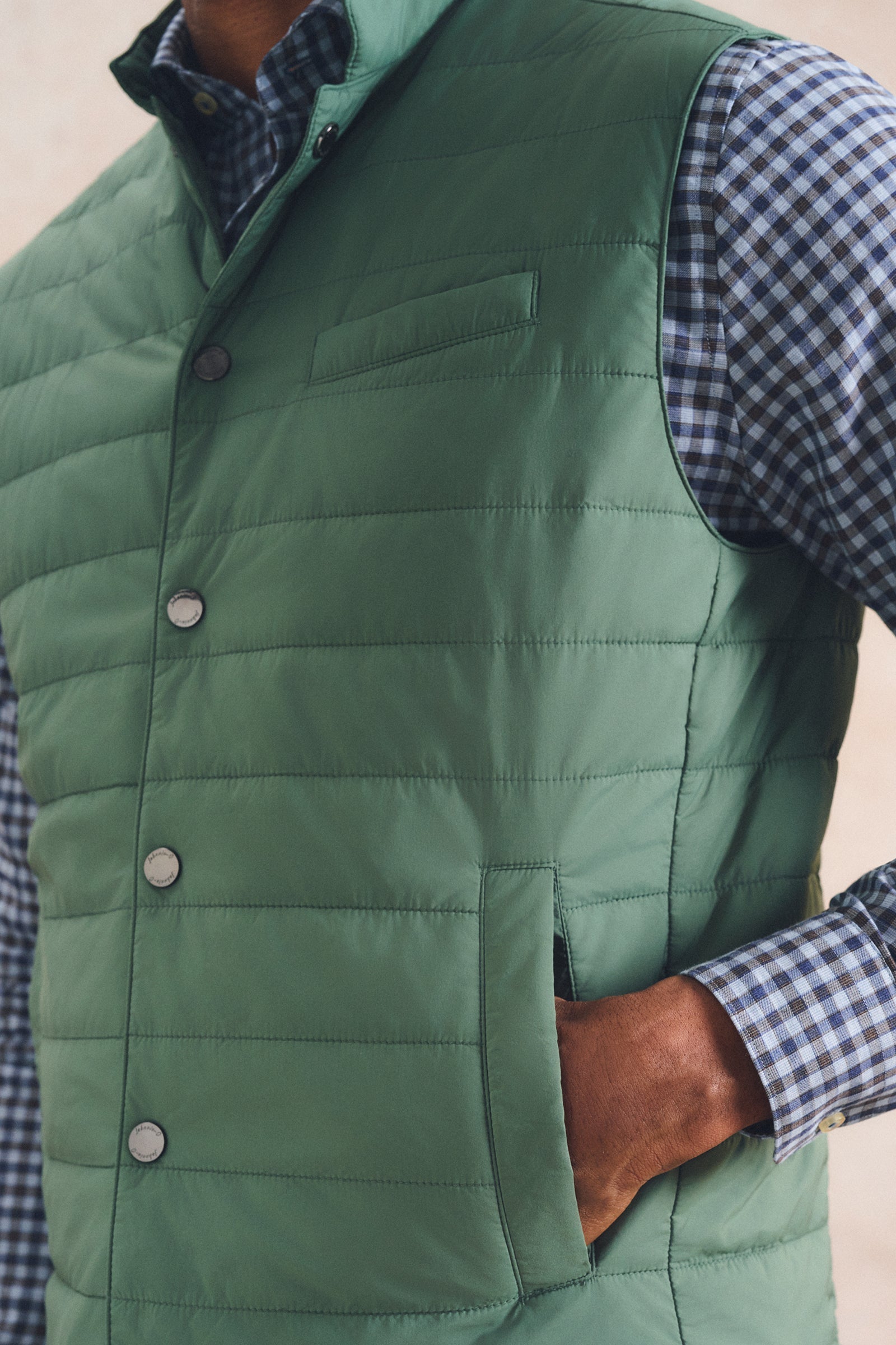 A close-up of a man wearing a sage green quilted puffer vest over a blue and white gingham check shirt, with his hand in his pocket.