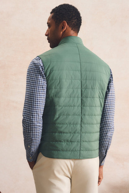 A man shown from the back wearing a sage green quilted puffer vest over a blue and white gingham shirt, paired with cream trousers, against a textured beige wall.