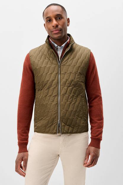 A man wearing a quilted olive green zip-up vest over a rust-colored long-sleeve shirt and cream pants, photographed against a light gray studio background.