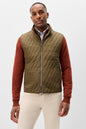 A man wearing a quilted olive green zip-up vest over a rust-colored long-sleeve shirt and cream pants, photographed against a light gray studio background.