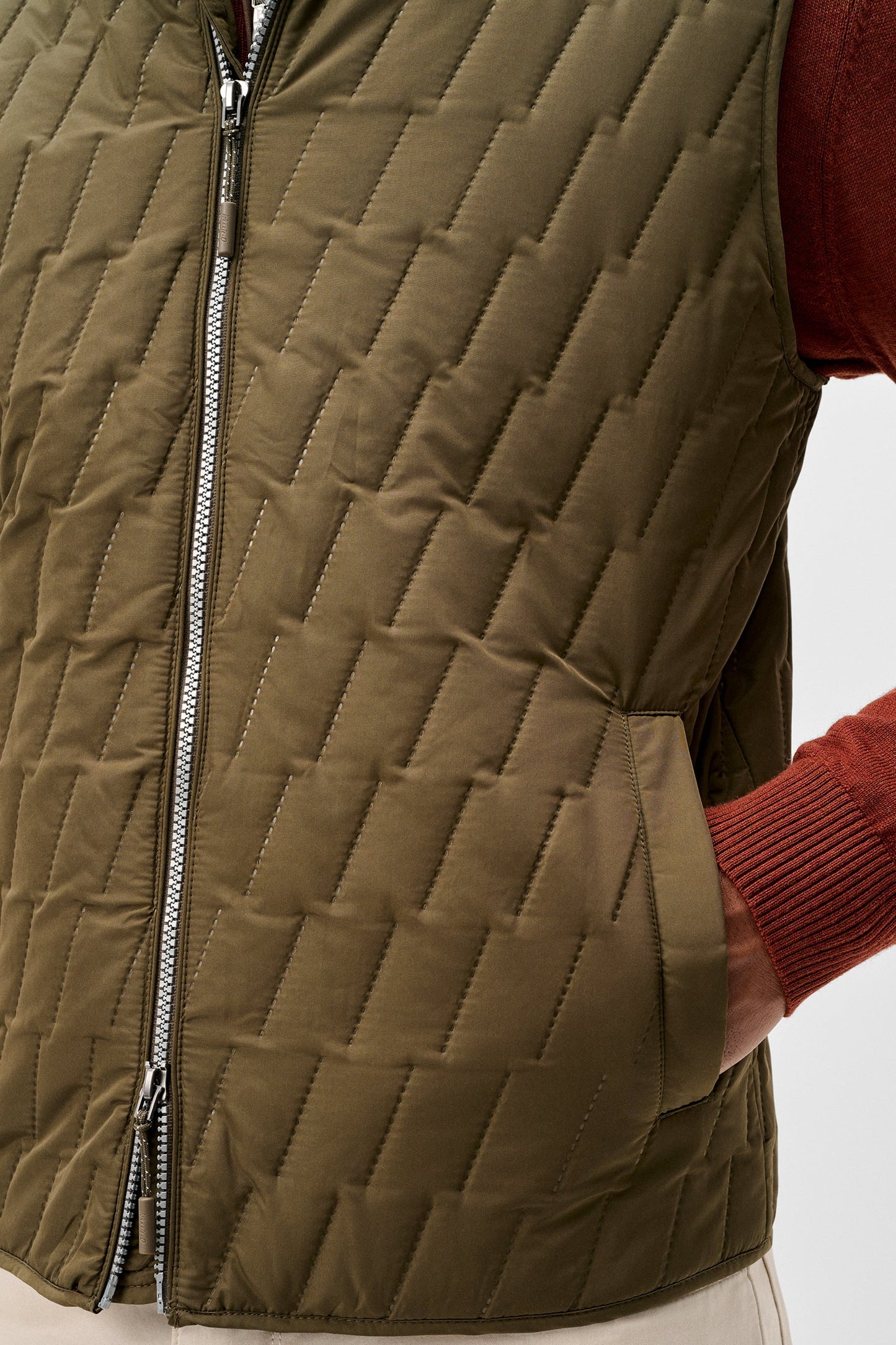 Close-up of a quilted olive green puffer vest worn over a rust-red ribbed sweater, with a silver zipper and side pockets, against a light gray background.