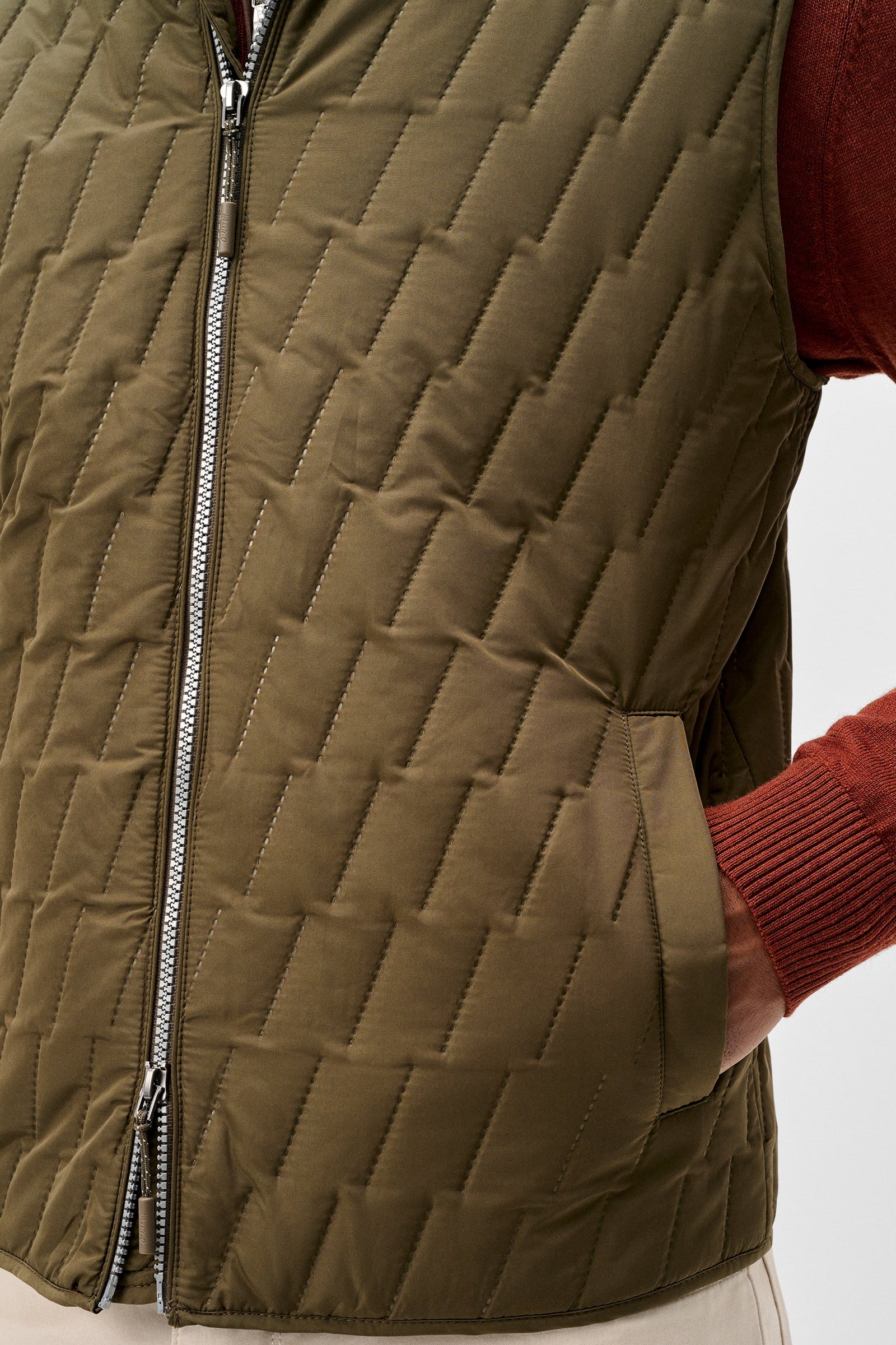Close-up of a quilted olive green puffer vest worn over a rust-red ribbed sweater, with a silver zipper and side pockets, against a light gray background.