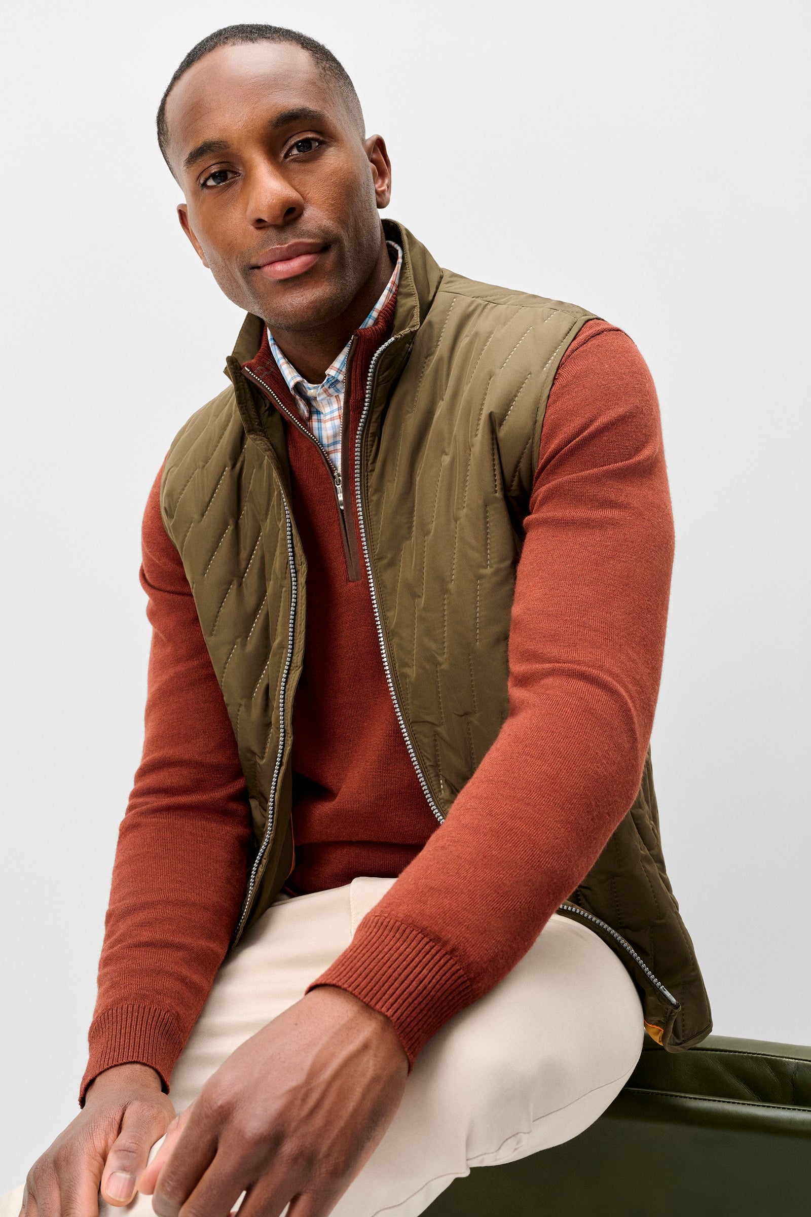 A man wearing a quilted olive green vest over a rust-orange long-sleeve sweater, cream trousers, and a patterned collared shirt, seated against a light gray studio background.