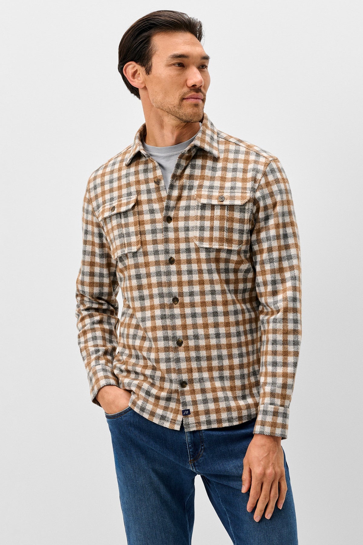 A man wearing a brown and beige plaid button-up shirt over a gray t-shirt and blue jeans, standing against a light gray studio background.