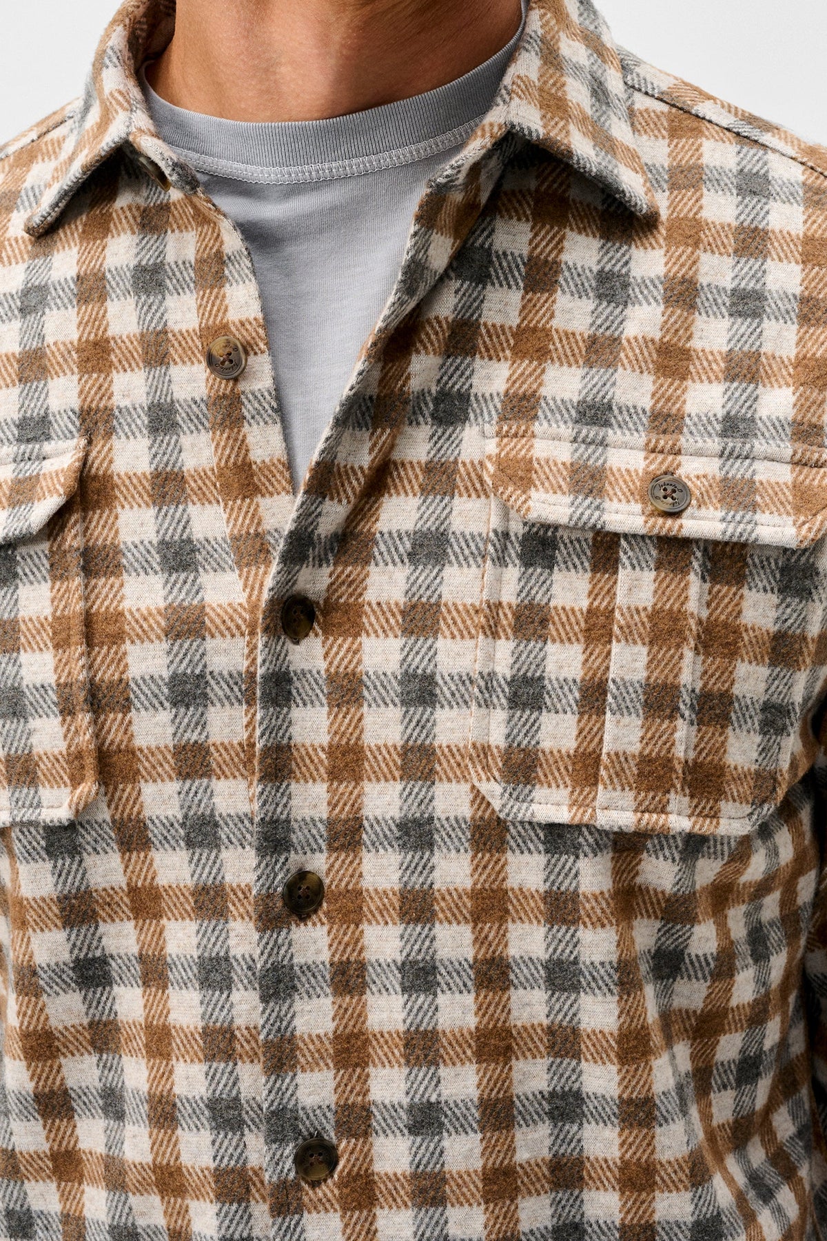 A close-up of a person wearing a brown, beige, and gray plaid button-up flannel shirt over a light gray t-shirt.