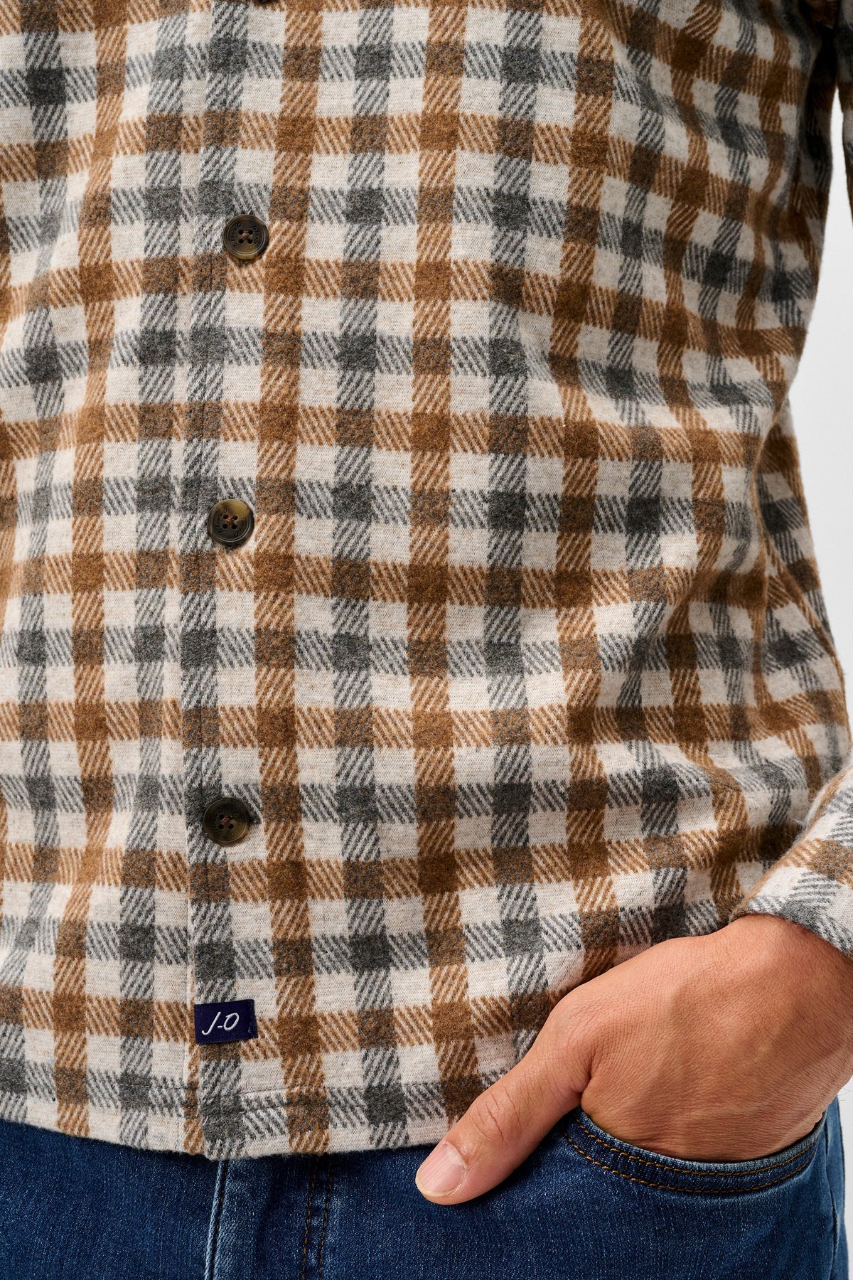Close-up of a person wearing a brown, beige, and gray plaid button-up shirt with a hand in a blue jeans pocket, against a light gray background.