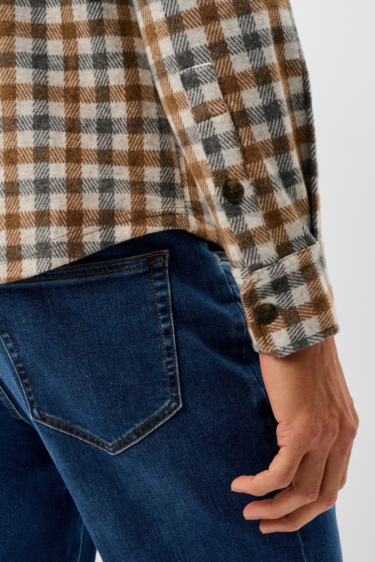 Close-up of a person wearing a brown, beige, and gray plaid flannel shirt tucked into dark blue jeans, showing the back pocket and hand.