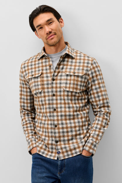 A man wearing a brown and beige plaid button-down flannel shirt over a gray t-shirt and blue jeans, standing against a light gray studio background.