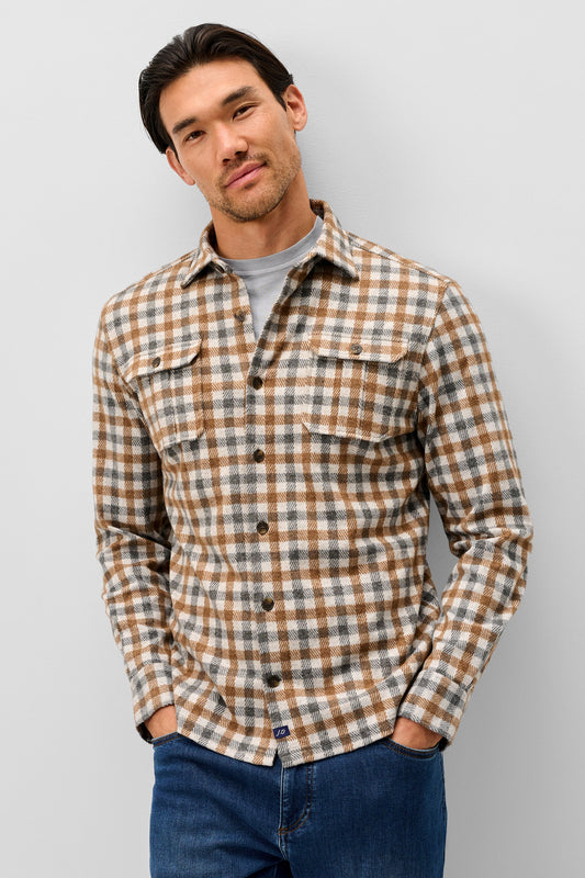 A man wearing a brown and beige plaid button-down flannel shirt over a gray t-shirt and blue jeans, standing against a light gray studio background.