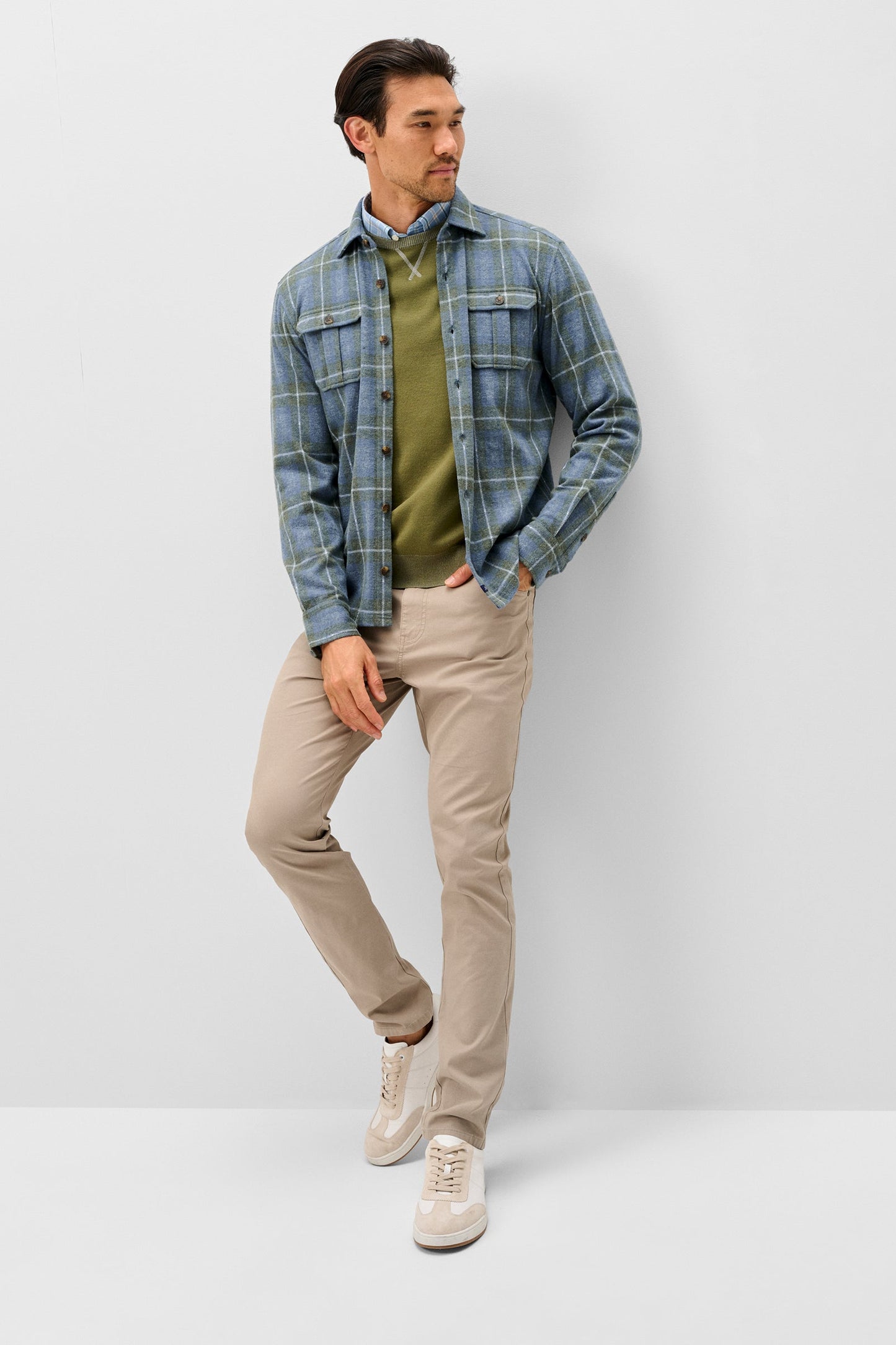 A man modeling a blue and green plaid flannel shirt over an olive green sweater, paired with beige trousers and white sneakers, standing against a light gray studio background.