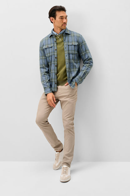 A man modeling a blue and green plaid flannel shirt over an olive green sweater, paired with beige trousers and white sneakers, standing against a light gray studio background.