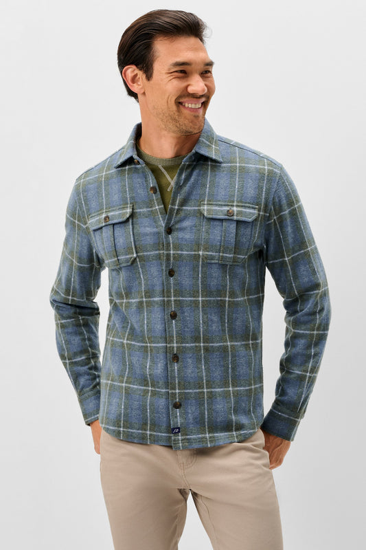 A man wearing a blue and green plaid button-down flannel shirt over an olive green t-shirt, paired with beige pants, photographed against a light gray studio background.