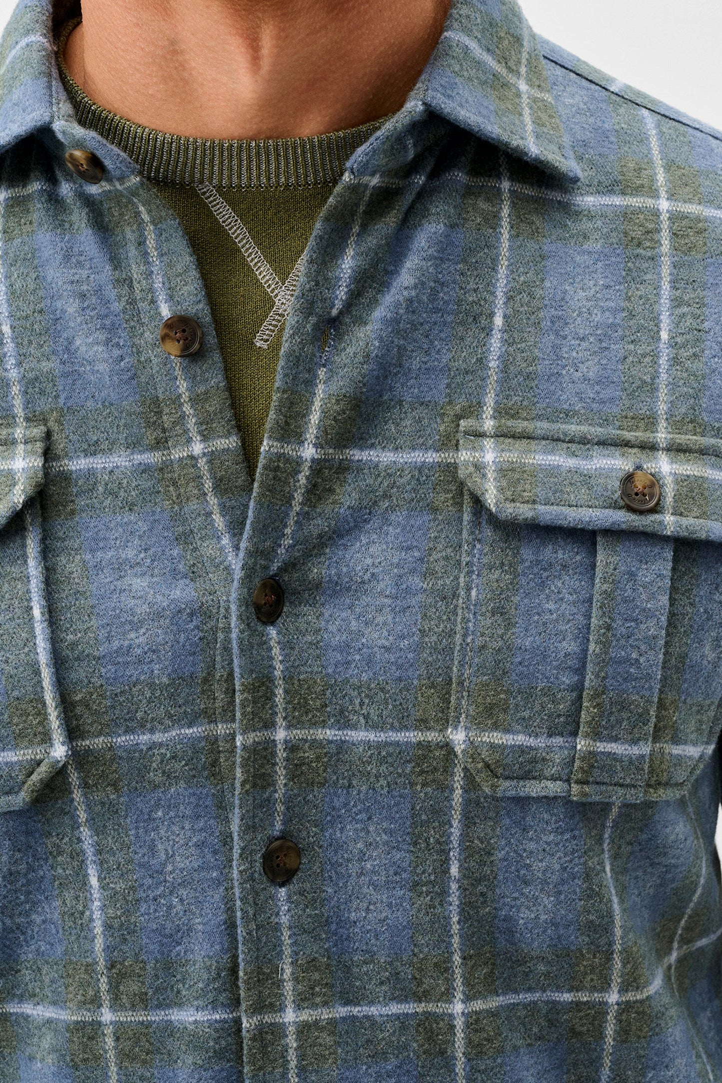Close-up of a person wearing a blue and green plaid button-up shirt over an olive green sweater, showing the collar, chest pocket, and buttons.